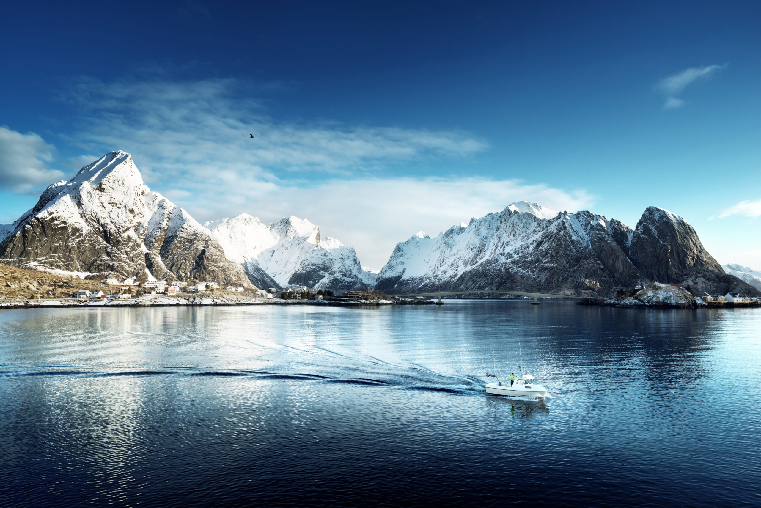 Bilde i farger som viser fjell i Lofoten med snø på toppen, en brygge i bakgrunnen og en båt som kjører på vannet