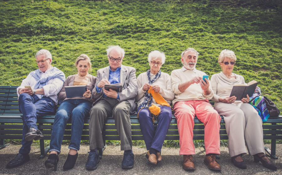Bildet viser en gruppe eldre mennesker på en benk i en park. Noen leser på mobilen, en leser bok, en strikker.