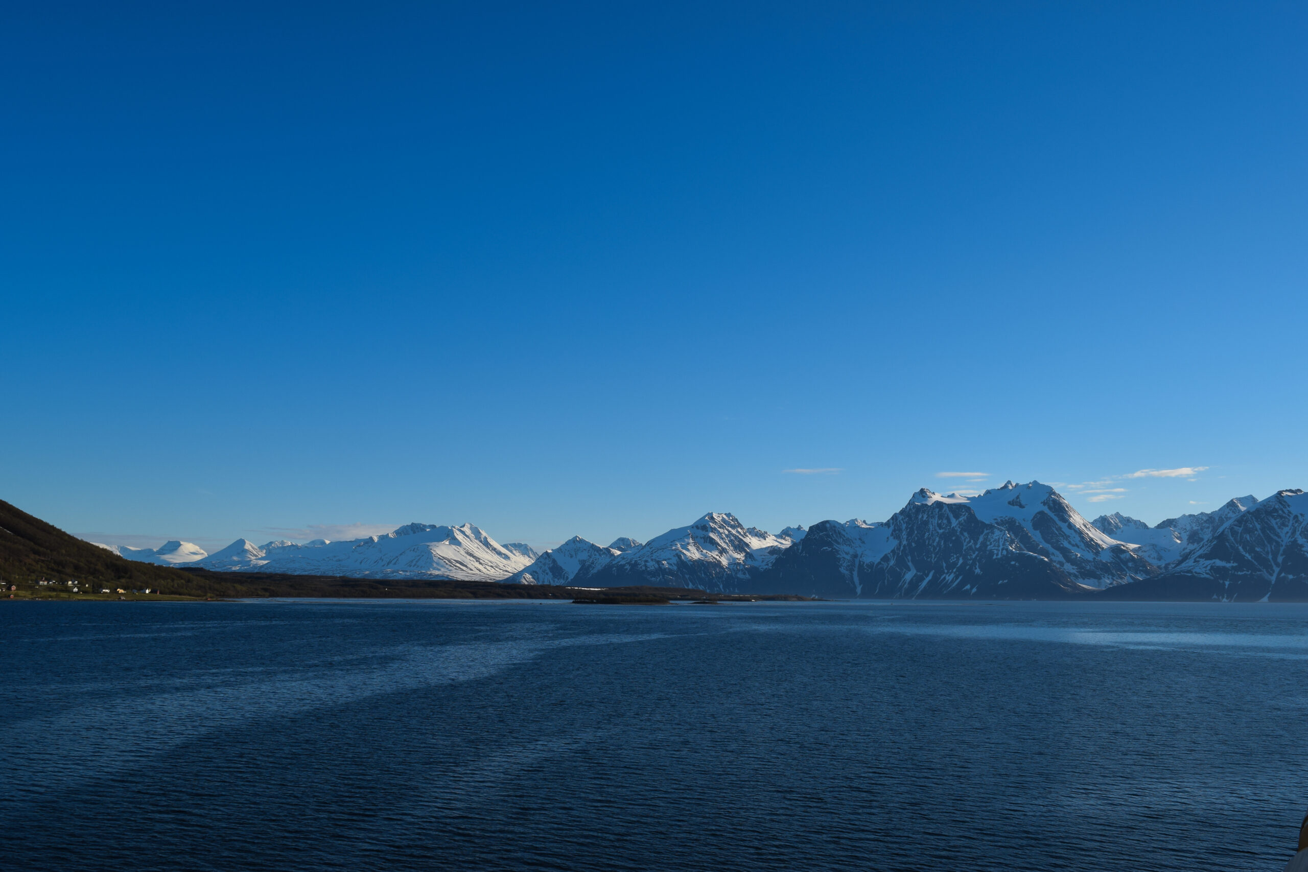 Bilde av fjell med snø langs kysten i Norge. Blå himmel og mørkeblått, stille hav.